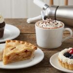 A delicious assortment of desserts on a wooden table: a cupcake, hot chocolate with whipped cream, apple pie slice, and fruit tart. A whipped cream dispenser is nearby.