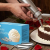 Hands using a cream charger to decorate a chocolate cake with whipped cream. Box of cream chargers nearby, red rose petals scattered on a wooden table.