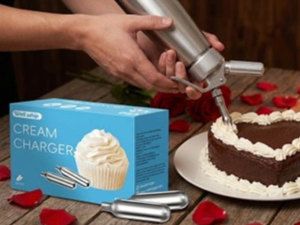 Hands using a cream charger to decorate a chocolate cake with whipped cream. Box of cream chargers nearby, red rose petals scattered on a wooden table.