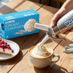 A hand uses a Wellwhip cream whipper on a cappuccino, producing swirls of whipped cream. Nearby, a cheesecake slice with berries and a box of cream chargers.