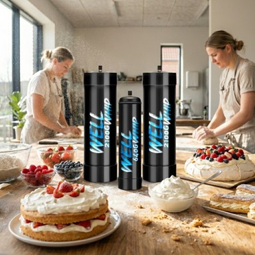 Two people baking in a bright kitchen, surrounded by cakes and berries. Three tall black canisters labeled "WELL WHIP" stand prominently on the table. The scene conveys a joyful and collaborative atmosphere.