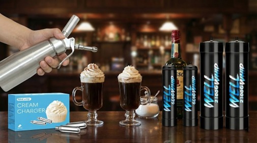 A hand holds a cream whipper above two coffee glasses topped with whipped cream. Nearby are cream charger boxes, a bottle of liqueur, and nitrogen canisters. The scene is set in a cozy bar with a warm ambience.