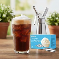 A glass of iced coffee topped with whipped cream sits on a wooden table beside a cream charger and dispenser, with potted plants in the background.