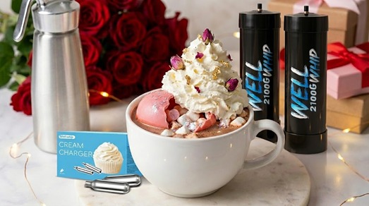 A whipped cream-topped hot chocolate with marshmallows in a white mug, surrounded by cream chargers, a dispenser, red roses, and gift boxes, creating a festive, cozy atmosphere.