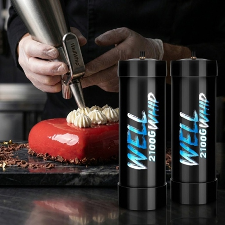 A chef in black attire pipes whipped cream onto a glossy red cake. Two black Well Whip cream chargers are in the foreground on a dark surface.