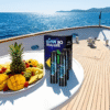 A yacht deck with a plate of tropical fruit, including pineapple and mango, sits beside a box of vaping products. The sea and distant coastline are visible under a clear sky.