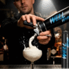 A bartender in a dimly-lit bar pours whipped cream into a cocktail glass using a black "Well Whip" canister, creating a splash. Candles in the background.