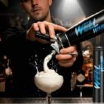 A bartender in a dimly-lit bar pours whipped cream into a cocktail glass using a black "Well Whip" canister, creating a splash. Candles in the background.