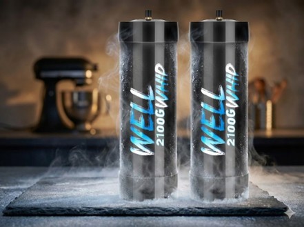 Two black metal canisters labeled "WELL 2100G WHIP" stand upright on a frosted surface with vapor around them, kitchen equipment blurred in the background.